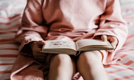 Foto de uma criança sentada na cama com um vestido rosa e comum livro aberto na mão.