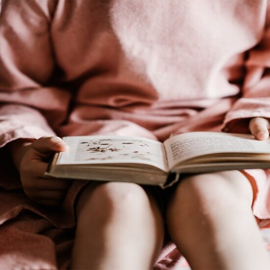 Foto de uma criança sentada na cama com um vestido rosa e comum livro aberto na mão.