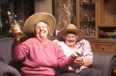 Giphy de duas senhoras amigas vestidas de rosa juntas sentadas no sofá comemorando com uma garrafa na mão cada uma.